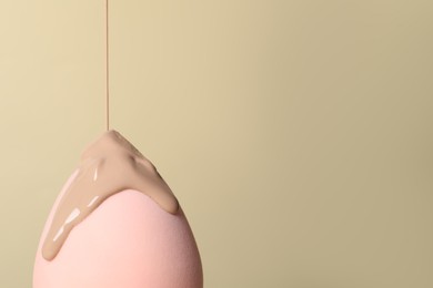Skin foundation pouring onto makeup sponge on beige background, space for text