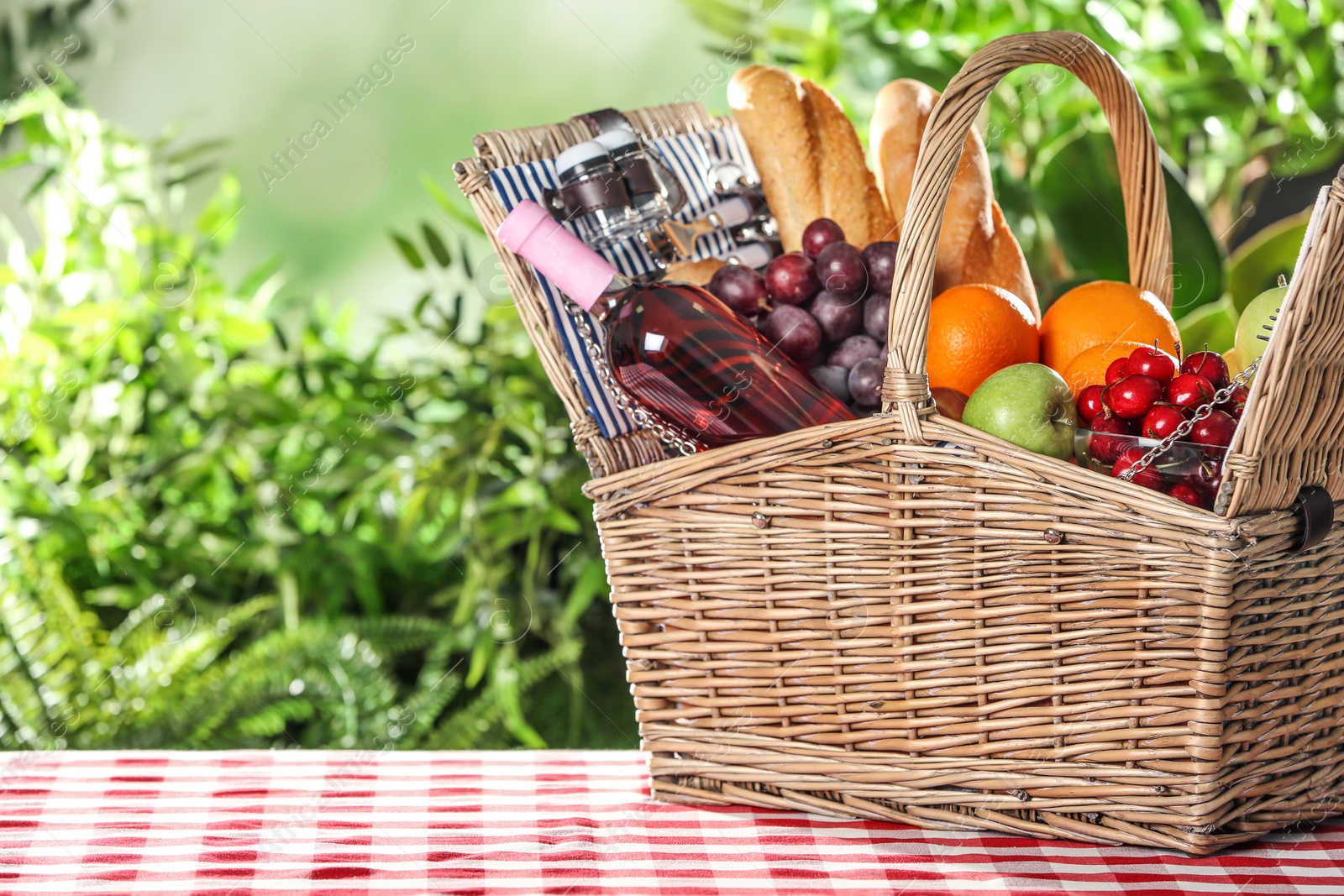 Photo of Wicker picnic basket with different products on checkered tablecloth against blurred background, space for text