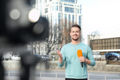 Photo of Young male journalist with microphone working on city street
