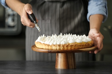 Photo of Woman toasting meringue on lemon pie with kitchen torch at table indoors, closeup