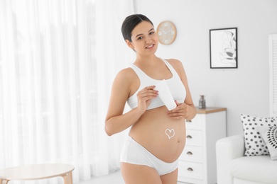 Photo of Pregnant woman with heart on her belly holding body cream at home