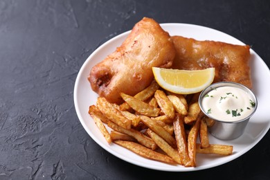Photo of Tasty fish, chips, lemon and sauce on black table, closeup. Space for text