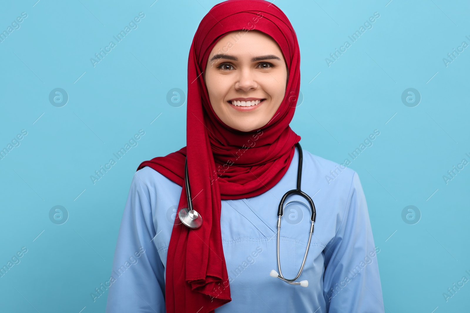 Photo of Muslim woman wearing hijab and medical uniform with stethoscope on light blue background