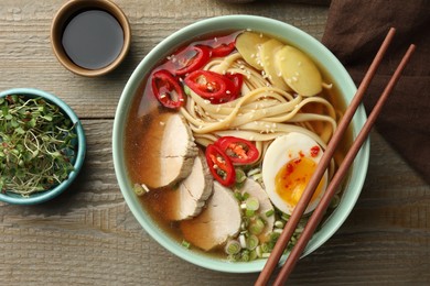 Photo of Delicious ramen in bowl, soy sauce and microgreens on wooden table, flat lay. Noodle soup
