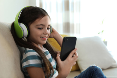 Photo of Cute little girl with headphones and smartphone listening to audiobook at home