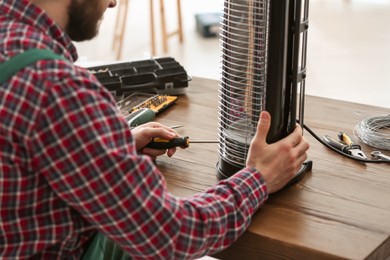Photo of Professional technician repairing electric halogen heater with screwdriver at table indoors, closeup