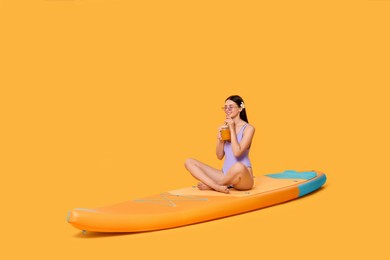 Happy woman with refreshing drink resting on SUP board against orange background