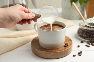Photo of Woman pouring milk into cup with coffee at white wooden table, closeup