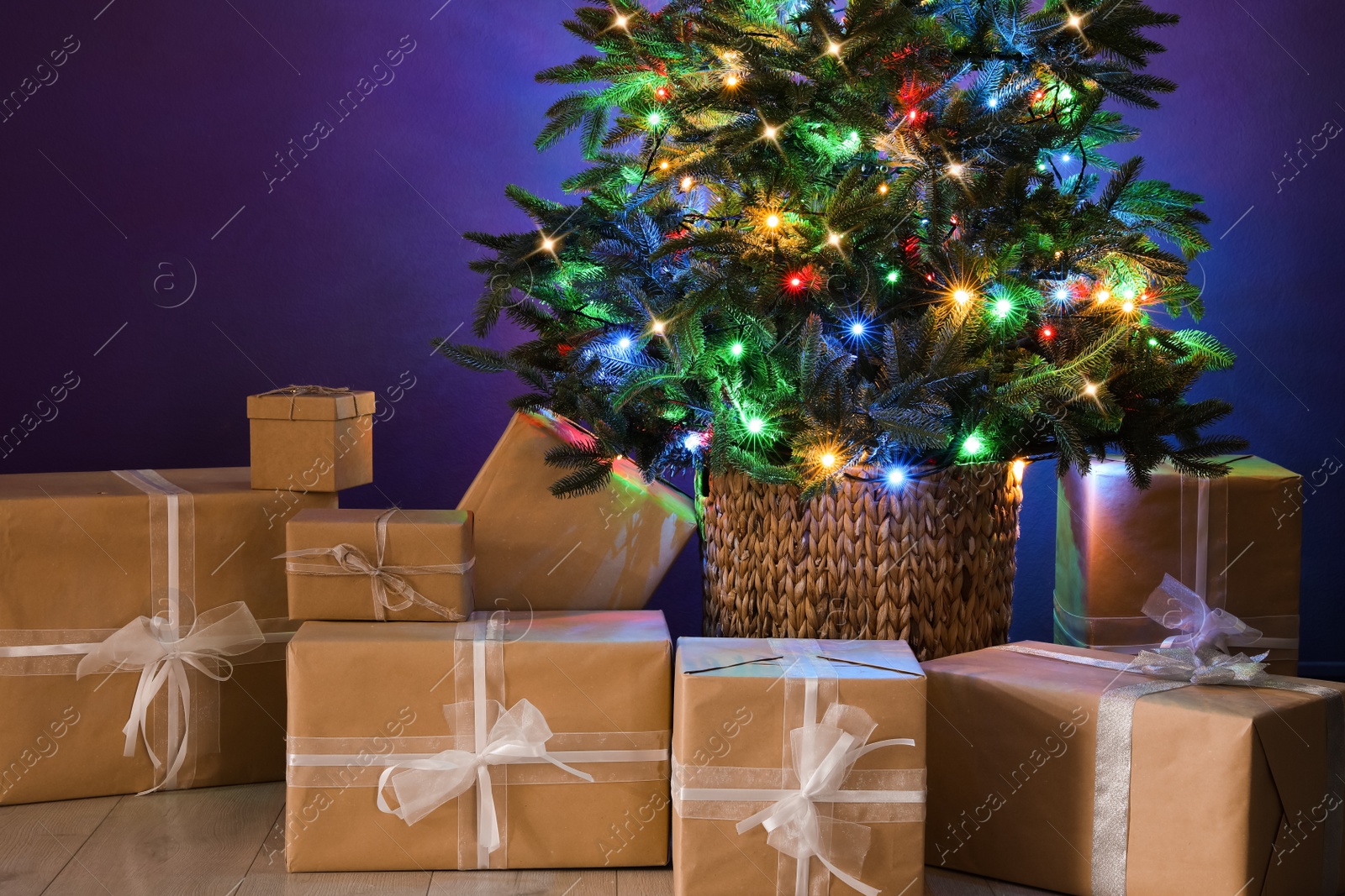 Photo of Beautiful Christmas tree with colorful lights and gift boxes near purple wall indoors