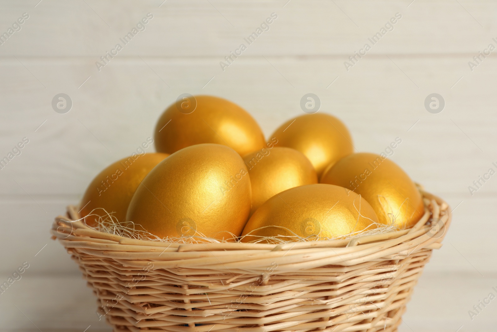Photo of Shiny golden eggs in wicker basket on light background, closeup
