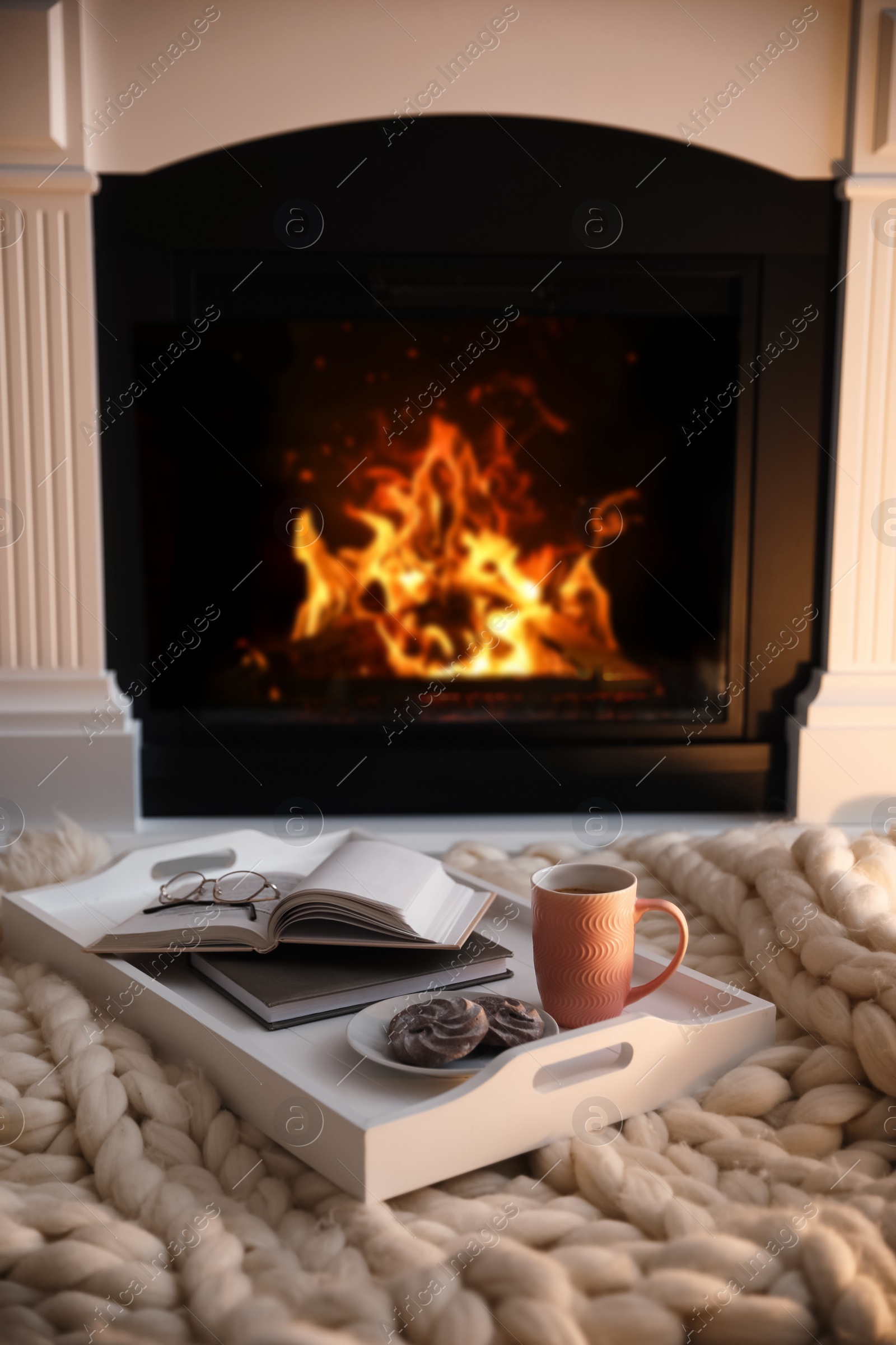 Photo of Cup of coffee, cookies and books on knitted blanket near burning fireplace indoors. Cozy atmosphere