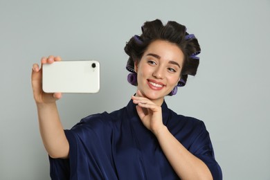 Photo of Happy young woman in silk bathrobe with hair curlers taking selfie on grey background