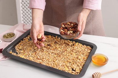 Photo of Making granola. Woman adding nuts onto baking tray with mixture of oat flakes and other ingredients at white table in kitchen, closeup