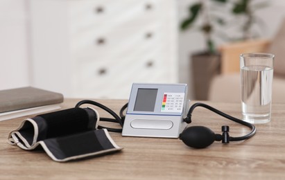 Photo of Modern blood pressure monitor and glass of water on wooden table indoors