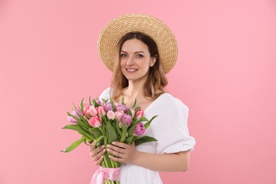 Happy young woman in straw hat holding bouquet of beautiful tulips on pink background