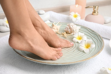 Photo of Woman soaking her feet in plate with water, flowers and seashells on white towel, closeup. Spa treatment