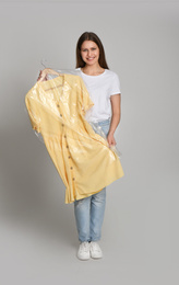 Young woman holding hanger with dress in plastic bag on light grey background. Dry-cleaning service
