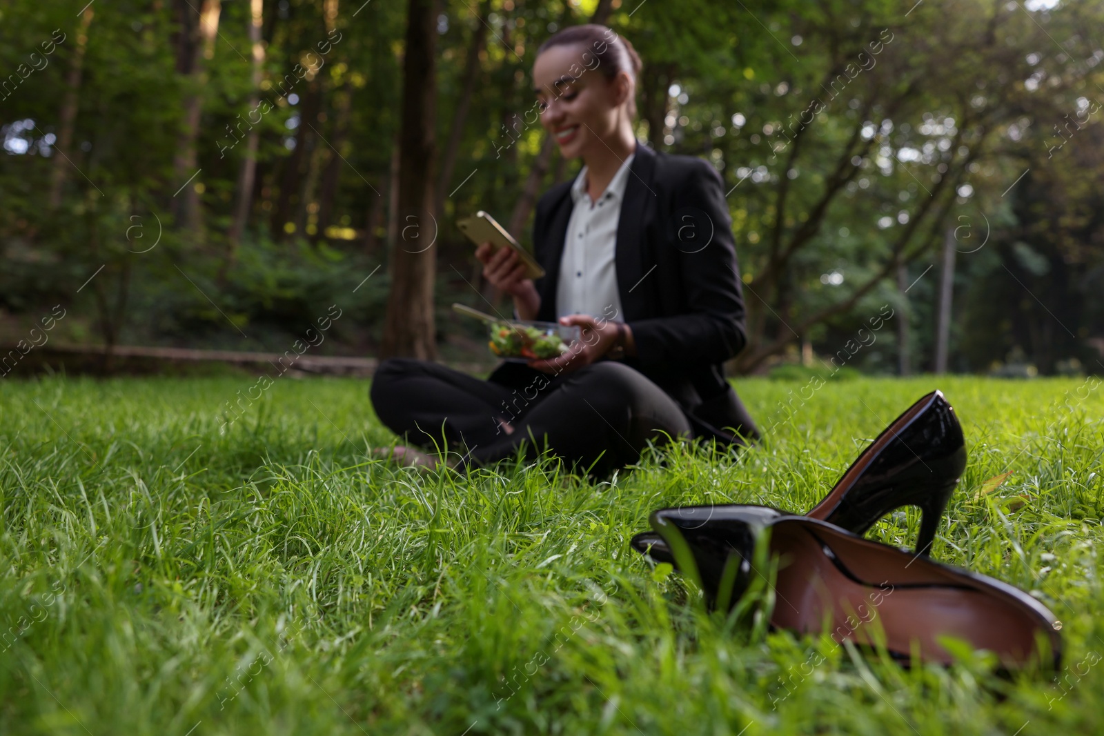 Photo of Lunch time. Businesswoman with container of salad using smartphone on green grass in park, focus on black shoes