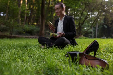 Photo of Lunch time. Businesswoman with container of salad using smartphone on green grass in park, focus on black shoes