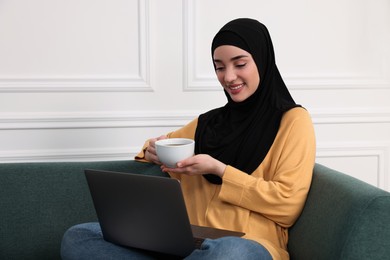 Muslim woman in hijab with cup of drink using laptop on sofa indoors