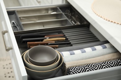 Open drawer of kitchen cabinet with different utensils, dishware and towels, closeup