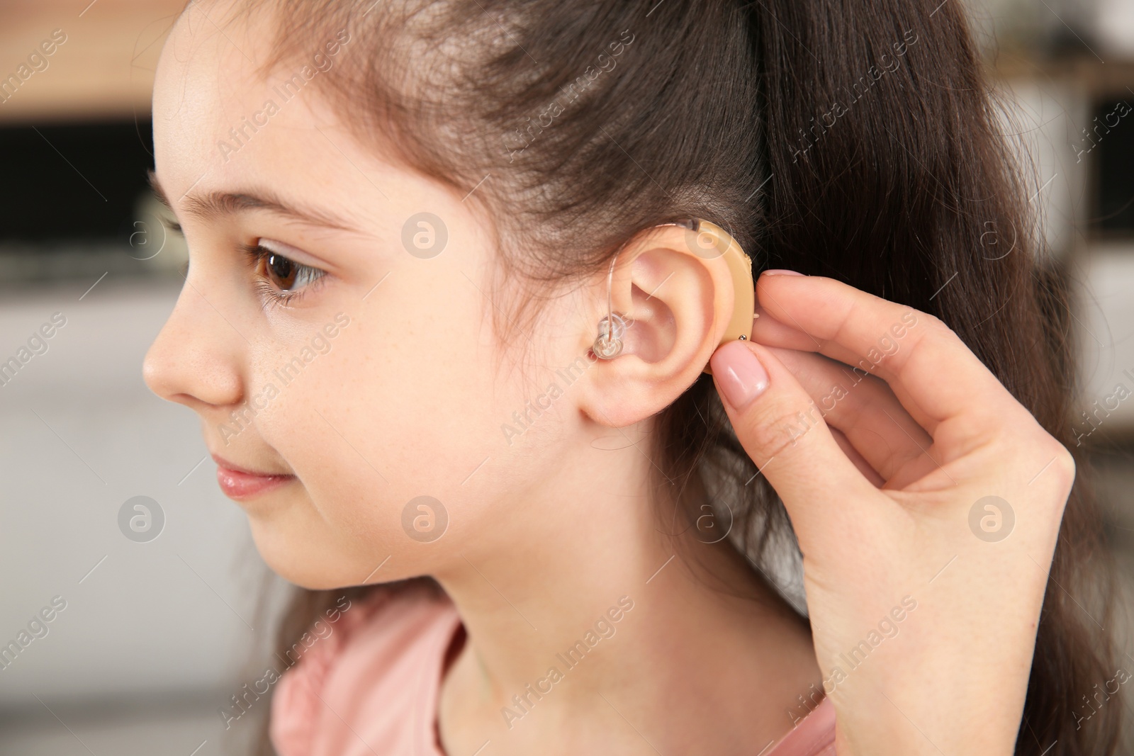 Photo of Young woman putting hearing aid in daughter's ear indoors, closeup