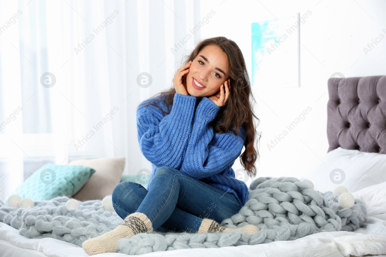 Photo of Young beautiful woman in warm sweater sitting on bed at home