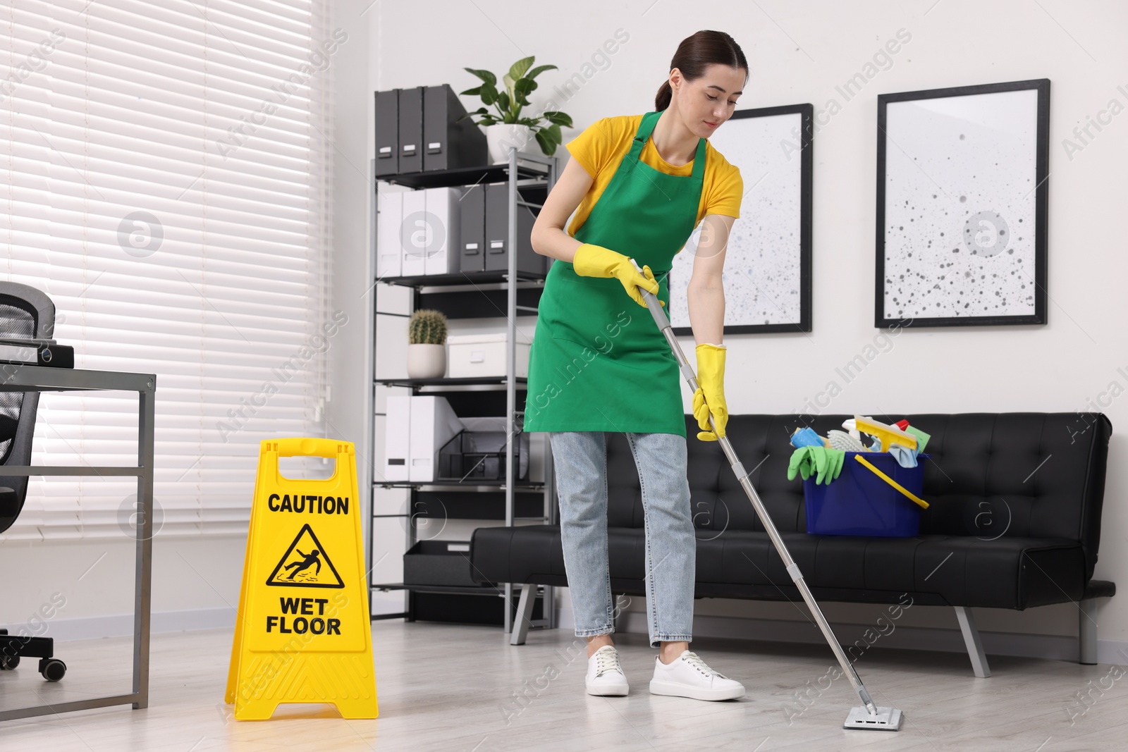 Photo of Cleaning service worker washing floor with mop. Bucket with supplies and wet floor sign in office