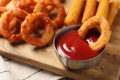 Photo of Tasty fried onion rings, cheese sticks and ketchup on wooden board, closeup
