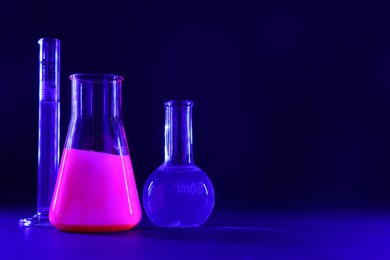 Photo of Laboratory glassware with luminous liquids on dark blue background, space for text