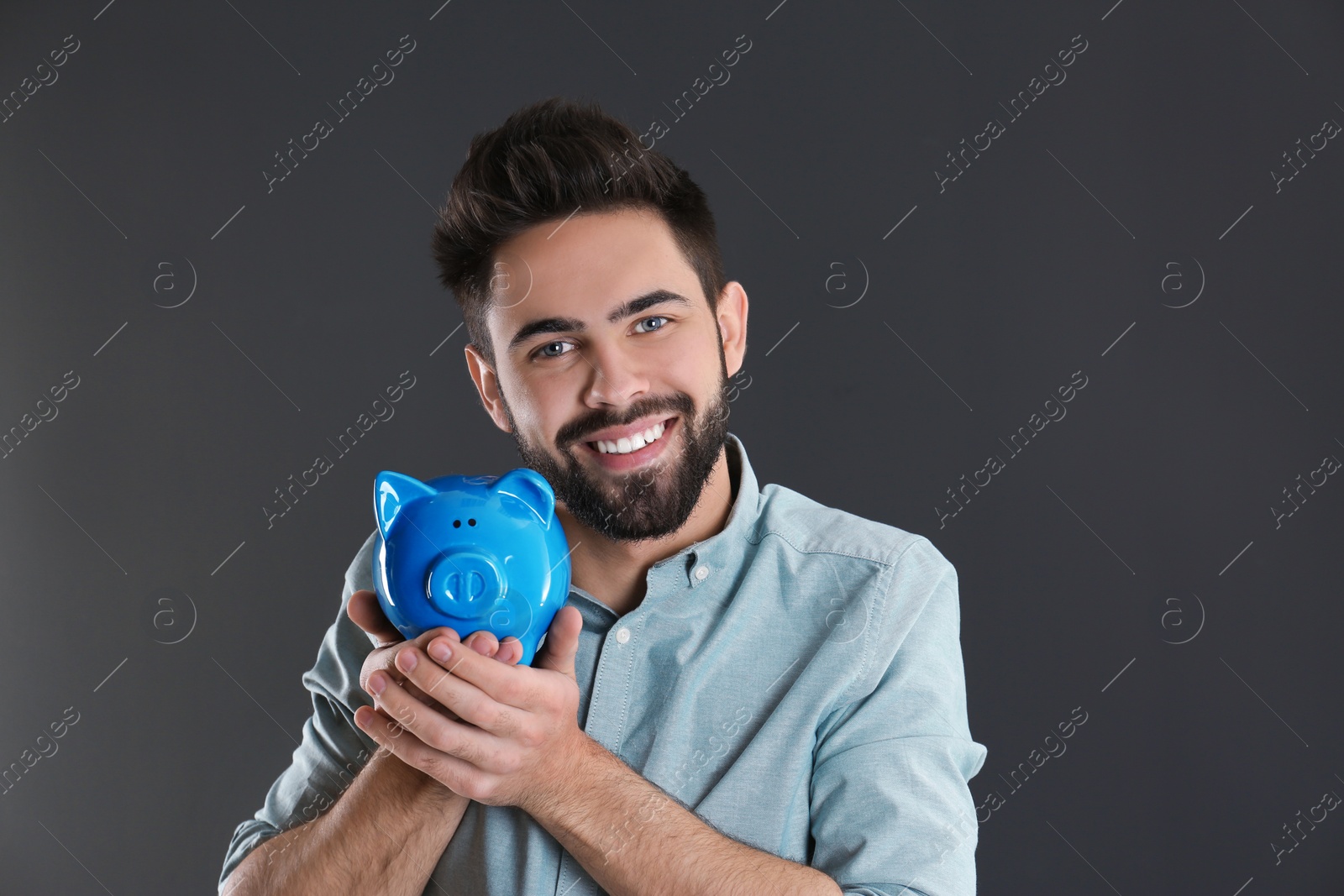 Photo of Portrait of happy young man with piggy bank on grey background. Money saving