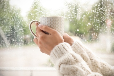 Photo of Young woman with cup of tea near window indoors on rainy day, closeup