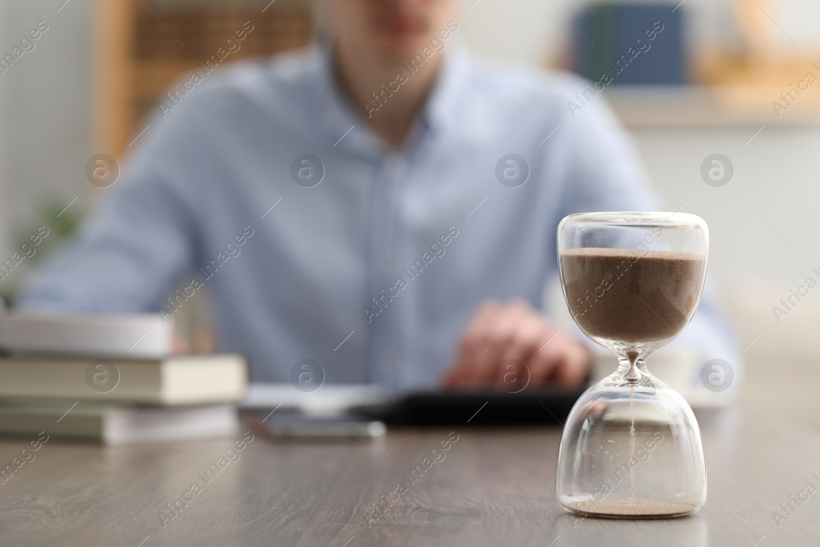 Photo of Hourglass with flowing sand on desk. Man using calculator indoors, selective focus