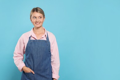 Beautiful young woman in denim apron on light blue background. Space for text