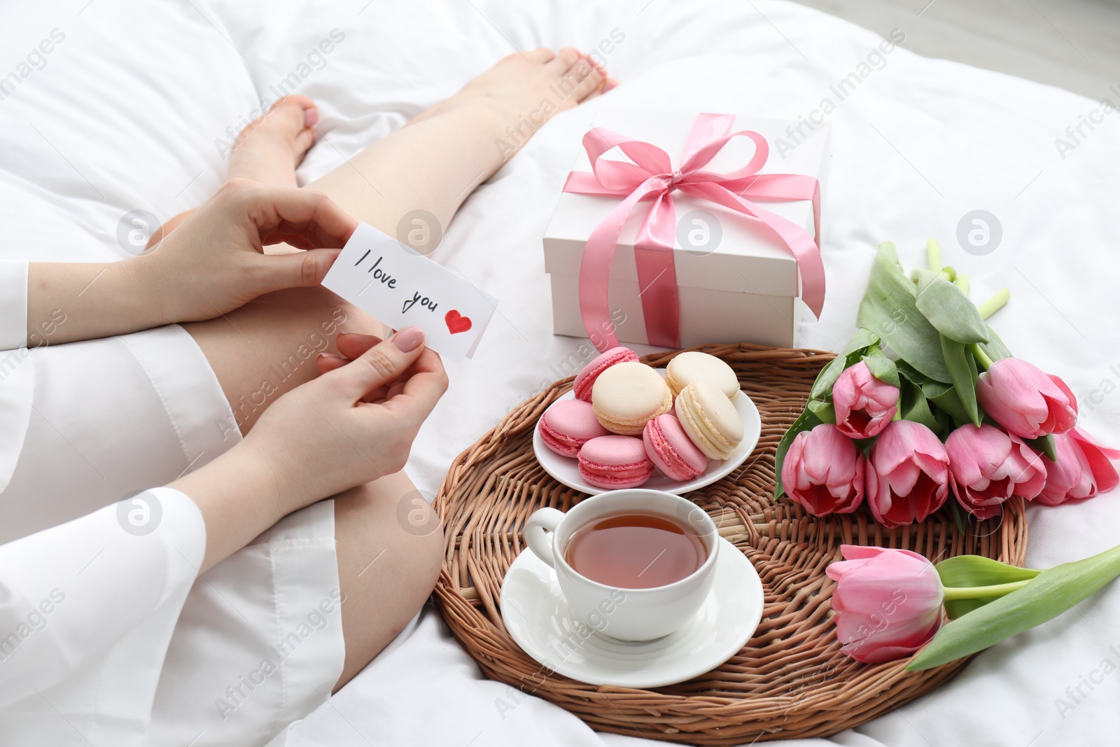 Photo of Tasty breakfast served in bed. Woman with I Love You card, macarons, tea, flowers and gift box at home, closeup