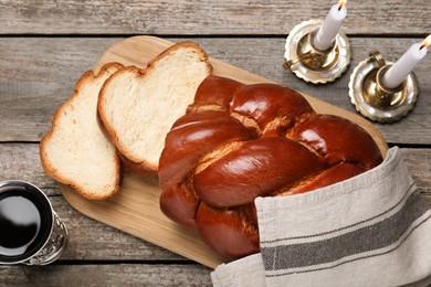 Photo of Cut homemade braided bread, goblet and candles on wooden table, flat lay. Traditional Shabbat challah