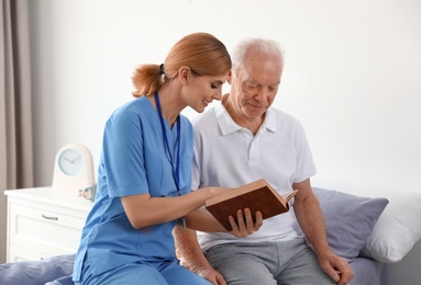 Photo of Nurse reading book to elderly man indoors. Medical assistance