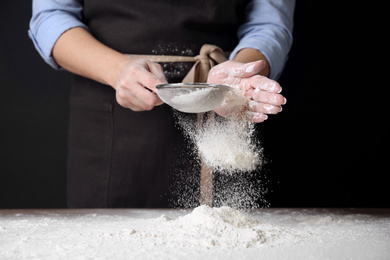 Photo of Woman sifting wheat flour at table against black background, closeup