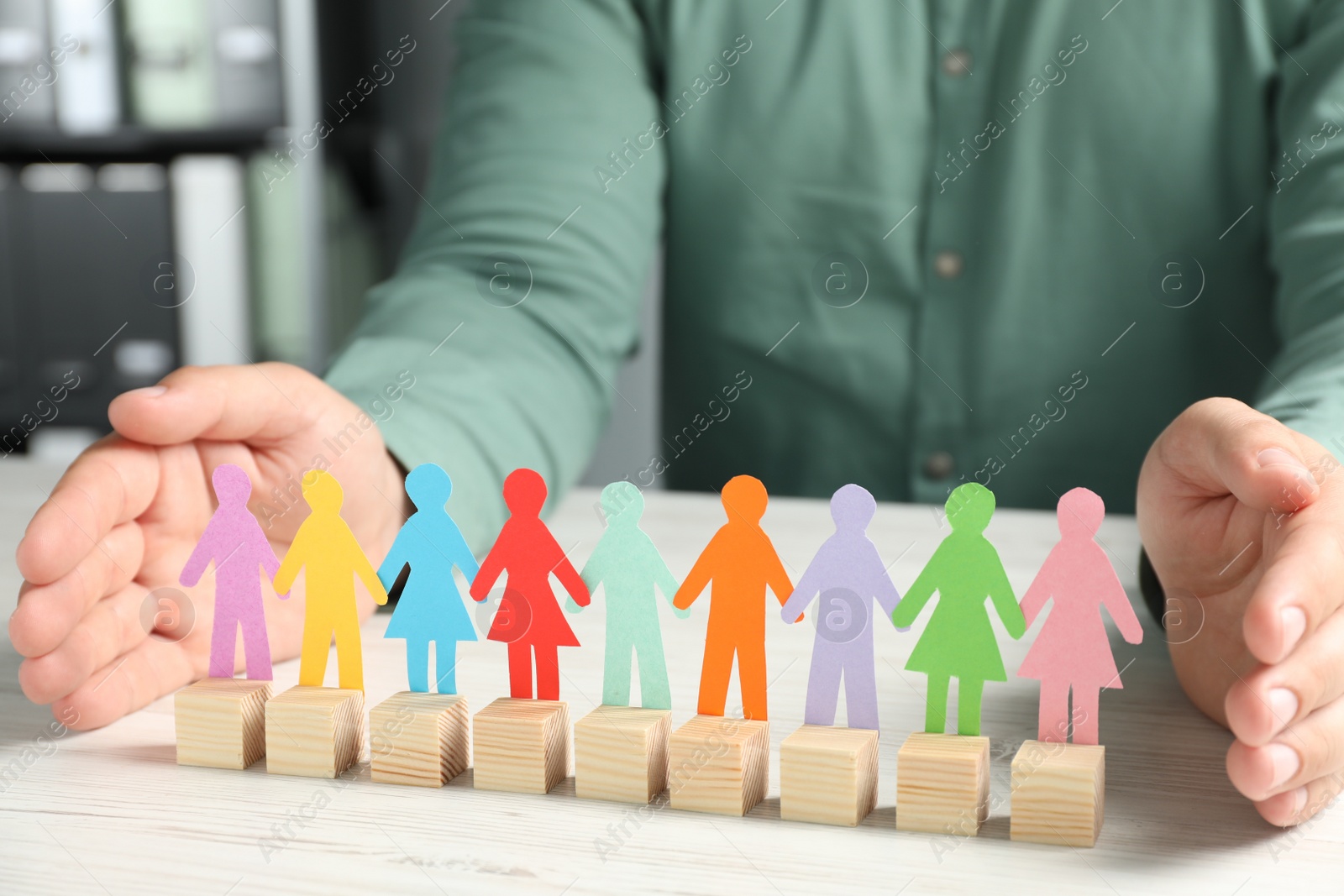 Photo of Man protecting paper human figures with wooden cubes at table indoors, closeup. Diversity and Inclusion concept