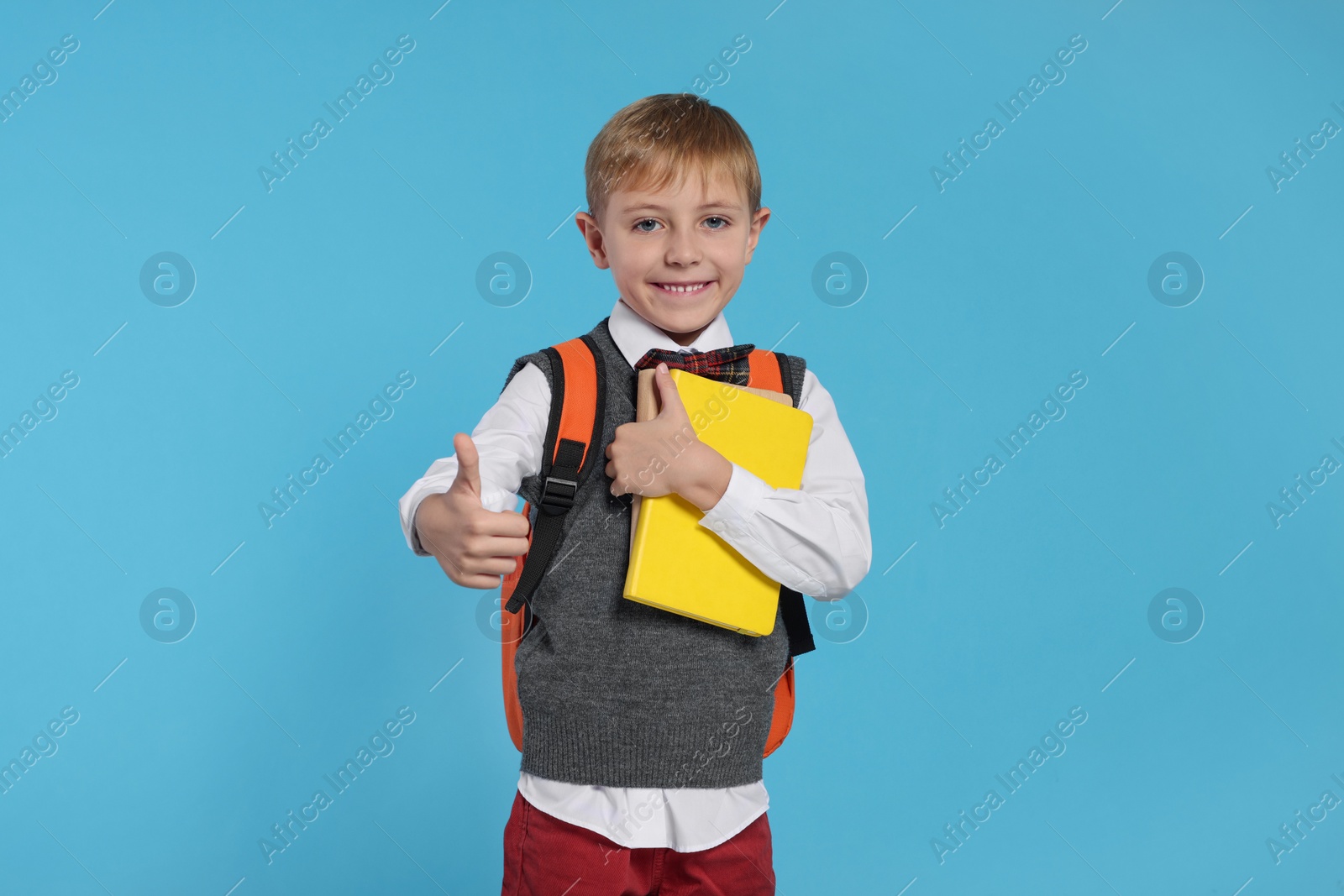 Photo of Happy schoolboy with backpack and books showing thumb up gesture on light blue background
