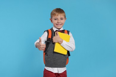 Happy schoolboy with backpack and books showing thumb up gesture on light blue background