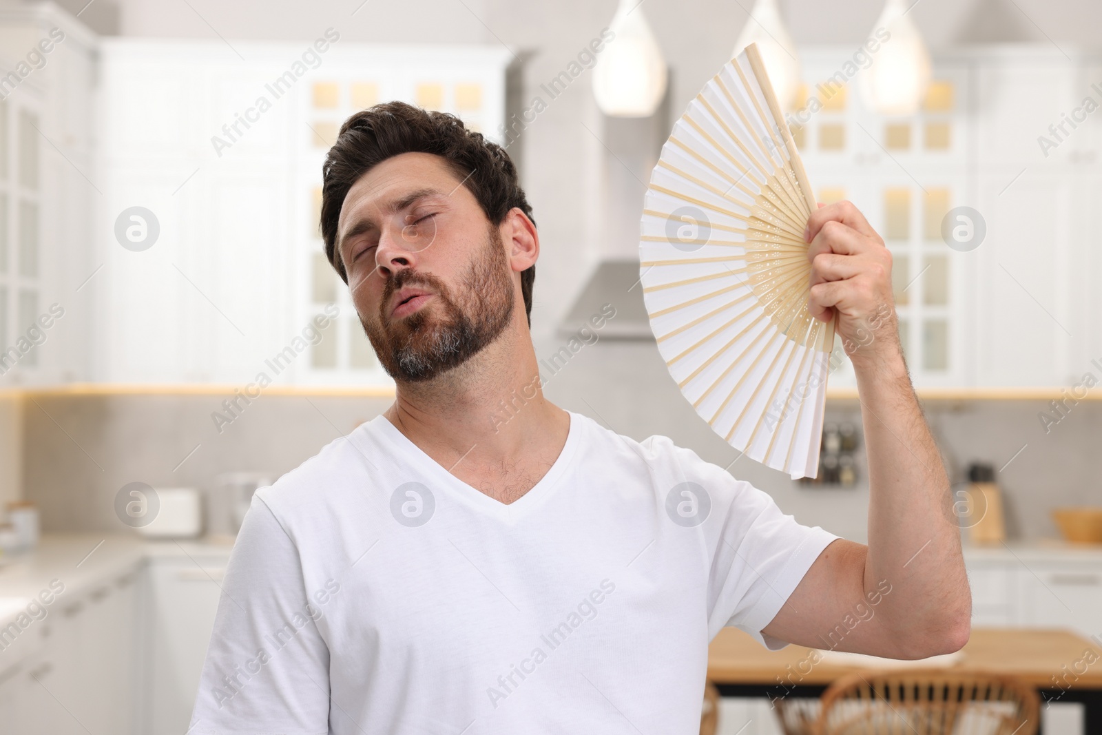 Photo of Bearded man waving white hand fan to cool himself in kitchen