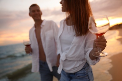 Photo of Couple drinking wine while walking near sea at sunset, focus on hand with glass