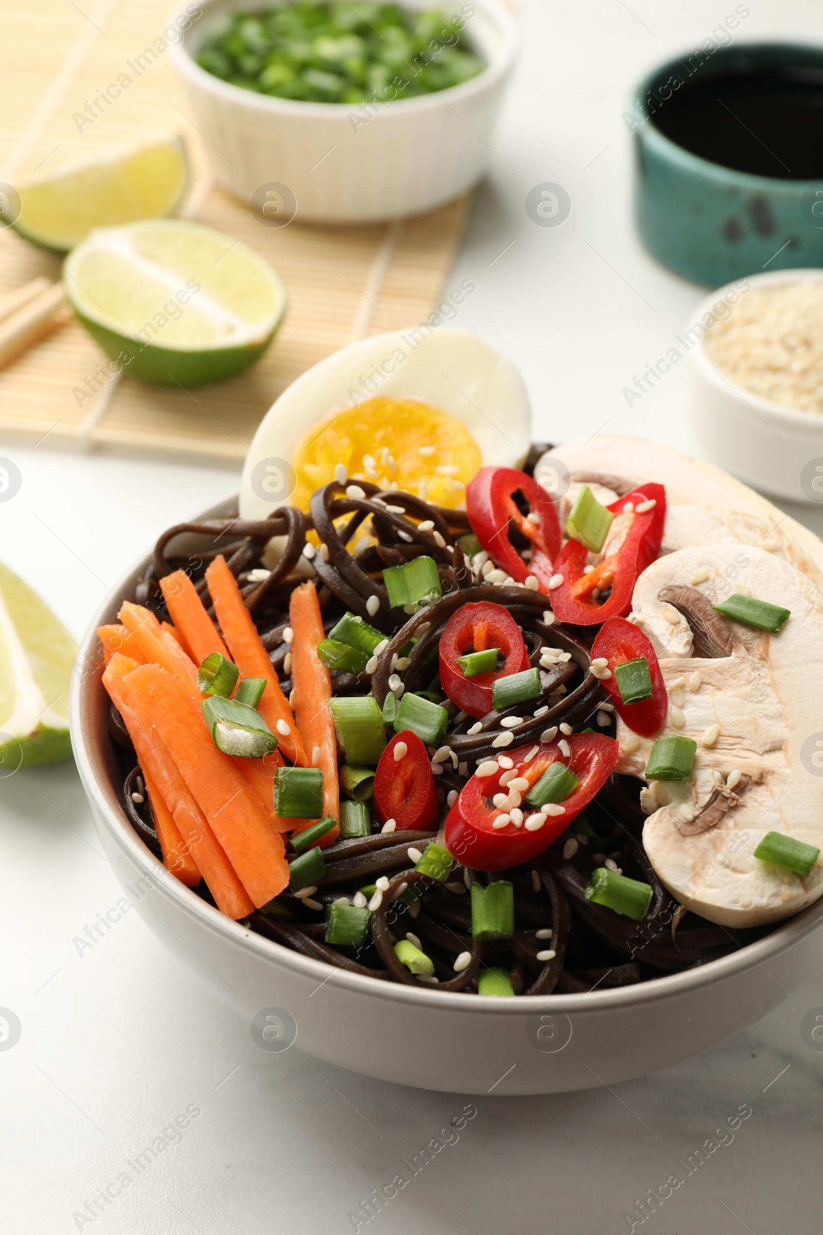 Photo of Tasty buckwheat noodles (soba) with chili pepper, egg, carrot and mushrooms in bowl on white marble table