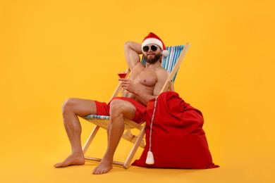 Photo of Muscular young man in Santa hat with deck chair, bag, sunglasses and cocktail on orange background
