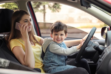 Mother with little son on knees driving car and talking by phone. Child in danger