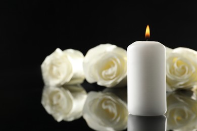 White roses and burning candle on black mirror surface in darkness, closeup with space for text. Funeral symbols