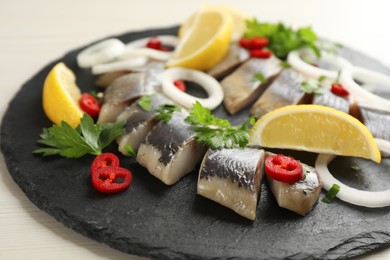 Photo of Slate plate with sliced salted herring fillet, parsley,  chili pepper, onion rings and lemon on light beige table, closeup