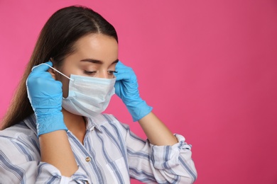 Woman in medical gloves putting on protective face mask against pink background. Space for text