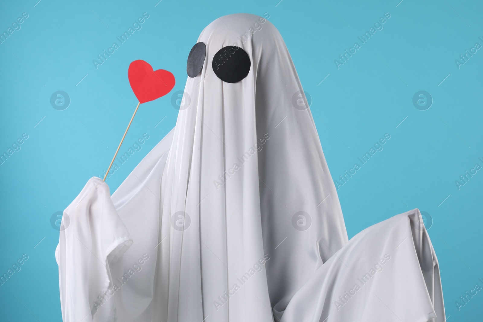 Photo of Cute ghost. Person covered with white sheet holding stick with red heart on light blue background